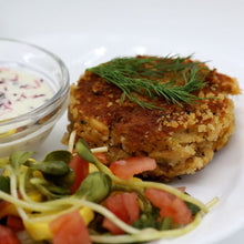 Load image into Gallery viewer, A golden-brown crab cake garnished with fresh dill sits on a white plate, accompanied by a colorful salad of diced tomatoes, yellow peppers, and greens. A small glass bowl of creamy dip with red flecks is in the background.